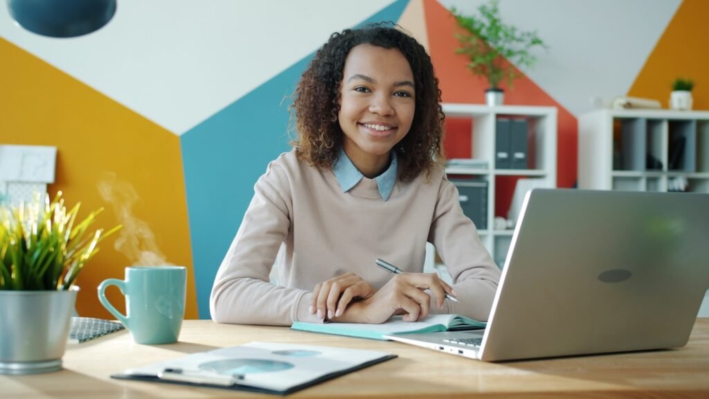 A young professional smiles while working at a desk with a laptop and a cup of coffee