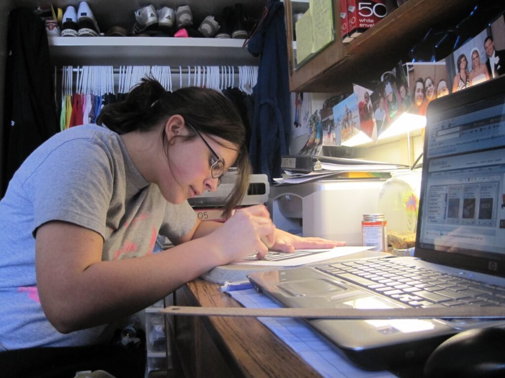 A student writing notes at a desk covered with open books and papers.