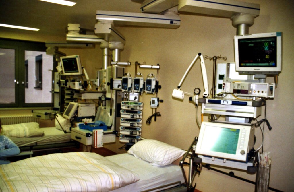 An empty intensive care bed with a made-up mattress, surrounded by a ventilator, infusion pumps and patient monitors.