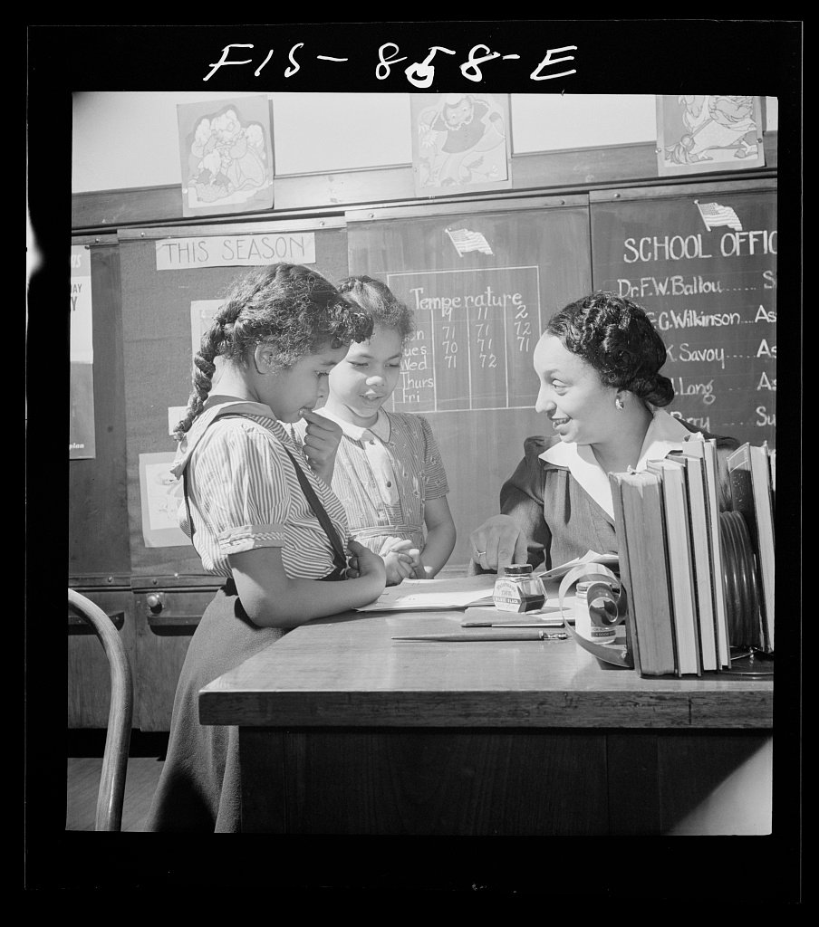 A teacher leaning over a desk to guide two pupils through their written work.
