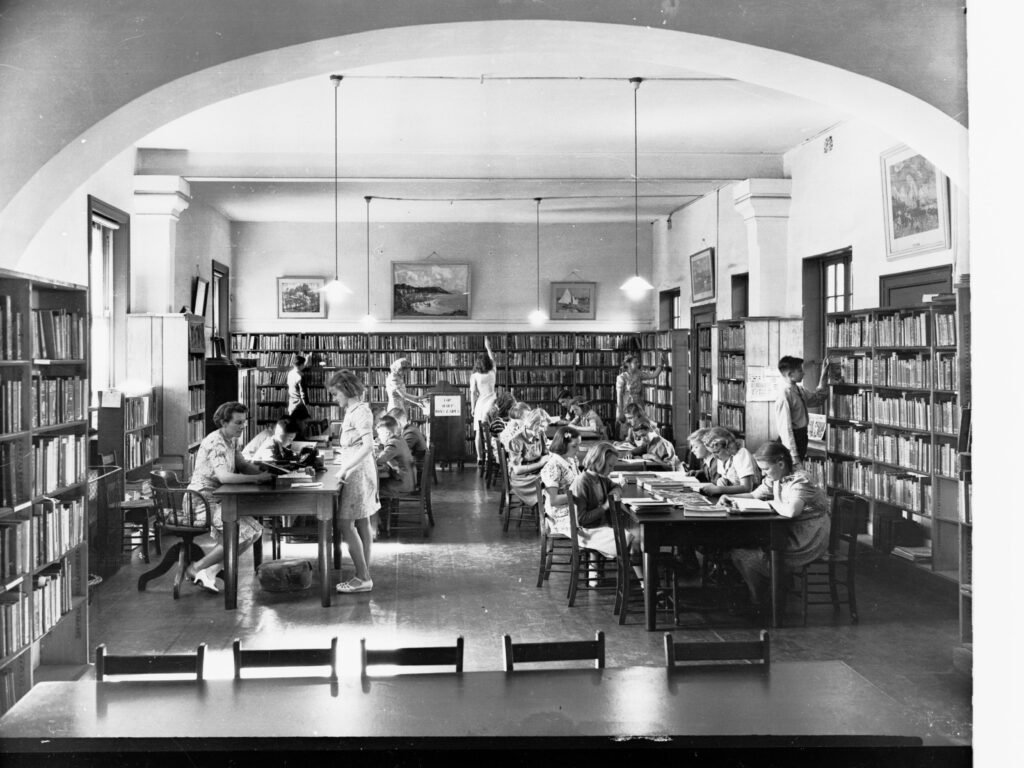 Students seated at tables in a school library, reading and taking notes.